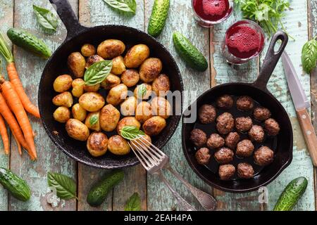 Skandinavische Küche. Gebratene Kartoffeln, Fleischbällchen und Preiselbeergetränk serviert mit Gemüse und Kräutern auf rustikalem blauen Tisch Overhead View Stockfoto