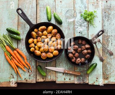 Nordisches Essen. Bratkartoffeln und Fleischbällchen in gusseisernen Pfannen mit rohen Bio-Karotten, Gurken und Kräutern auf schäbigen blauen Brettern Draufsicht Stockfoto