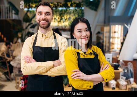 Porträt von zwei freundlichen Kellnern. Attraktive kaukasischen Kerl und schöne asiatische Mädchen in schwarzen Schürzen stehen mit Armen in einem Restaurant, Café oder Bar gekreuzt und lächelnd. Teamwork-Konzept Stockfoto