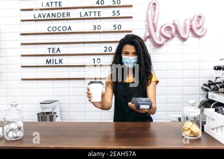 Eine freundliche afroamerikanische Kellnerin, die eine medizinische Maske trägt, die einem Besucher eine Papiertasse Kaffee und ein Terminal für kontaktlose Bezahlung gibt und im Coffee Shop steht Stockfoto