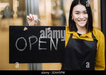 Wir sind offen. Unverschämtes zufriedene junge Frau, Kellnerin oder Inhaberin eines Cafés oder Restaurants mit schwarzer Schürze steht am Eingang mit einem Schild OFFEN und lächelnd. Konzept für kleine Unternehmen Stockfoto