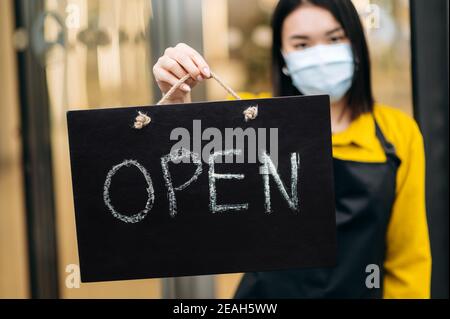 Schild IM Vordergrund GEÖFFNET. Entkokettete Kellnerin in einer schützenden medizinischen Maske auf ihrem Gesicht, hält Schild OFFEN am Eingang zu einem Restaurant oder Café. Konzept für kleine Unternehmen Stockfoto
