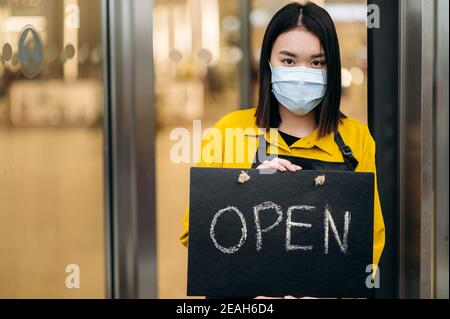 Porträt einer glücklichen Kellnerin, die am Eingang des Restaurants oder Cafés steht und eine medizinische Maske auf ihrem Gesicht trägt. Junge asiatische Frau zeigt offene Zeichen in ihrem kleinen Geschäft und lädt Kunden Stockfoto