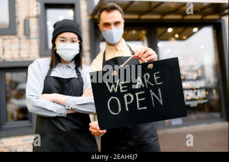 Schild IM Vordergrund GEÖFFNET. Zwei entkochte Kellner mit Schutzmasken, kaukasischer Mann und asiatische Frau, die vor einem Restaurant, Café oder Bar stehen und das Schild OFFEN zeigen, willkommen Stockfoto