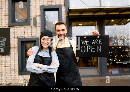 Wir sind OFFEN. Zwei multirassische freundliche Kellner in Uniform stehen vor einem Restaurant, Café oder Bar, zeigen Schild OFFEN, mit einem angenehmen Lächeln, Herzlich Willkommen. Teamwork-Konzept Stockfoto