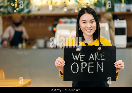 Wir sind offen. Portrait einer glücklichen hübschen asiatischen Kellnerin, die drinnen in einem Restaurant oder Café mit schwarzer Schürze steht, hält EIN Schild OFFEN und lächelt freundlich Stockfoto