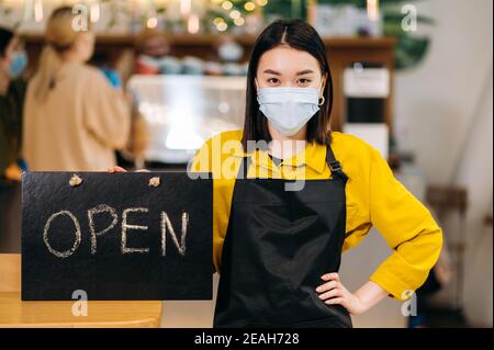 Herzlich Willkommen. Die junge asiatische Kellnerin steht im Innenbereich eines Restaurants oder Cafés mit einer schützenden medizinischen Maske und einer schwarzen Schürze und hält das Schild OFFEN. Unterstützung des Konzepts für kleine Unternehmen Stockfoto