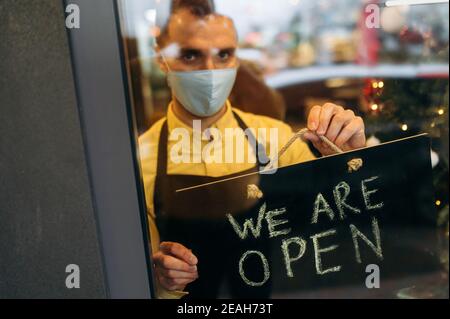 Herzlich Willkommen. Ein zufriedener kaukasischer Kellner oder Barista, der sein Schild OFFEN zeigt, während er am Eingang des Restaurants oder Cafés steht und eine medizinische Maske auf seinem Gesicht und eine schwarze Schürze trägt, lädt Gäste ein Stockfoto