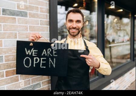 Willkommen, wir sind offen. Porträt des kaukasischen attraktiven männlichen Kellner im Freien in der Nähe eines Restaurants oder Cafés, zeigt einen Finger auf ein Schild OFFEN und freundliches Lächeln. Unterstützung des Konzepts für kleine Unternehmen Stockfoto