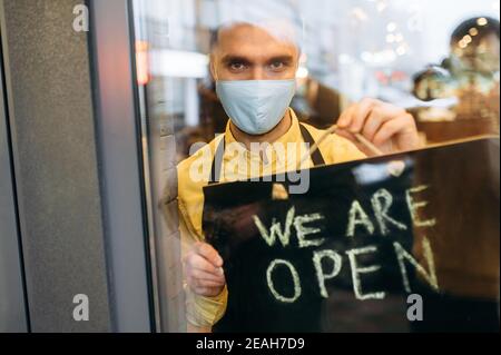Herzlich Willkommen. Ein zufriedener kaukasischer Kellner oder Barista, der sein Schild OFFEN zeigt, während er am Eingang des Restaurants oder Cafés steht und eine medizinische Maske auf seinem Gesicht und eine schwarze Schürze trägt, lädt Gäste ein Stockfoto
