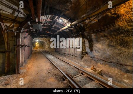 Tunnel des Bergbaus einer unterirdischen Mine. Viele Rohrleitungen an der Decke und Schienenbahn für Trolleys Stockfoto