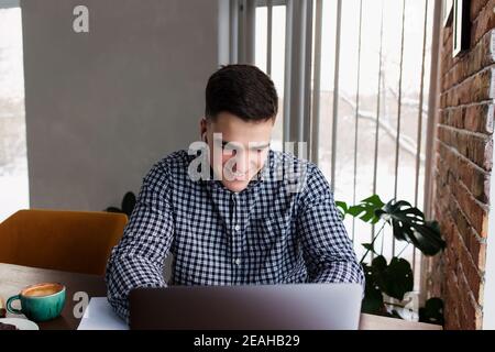 Lächelnder Mann, der am Laptop mit einer Tasse Kaffee zu Hause arbeitet. Unscharfer Hintergrund Stockfoto