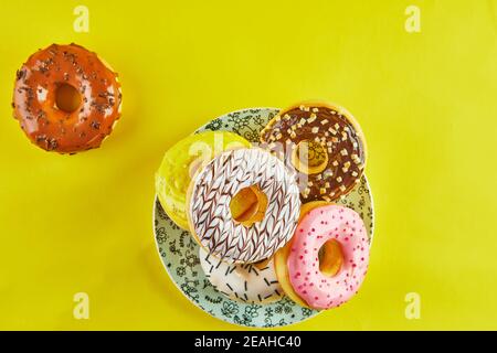 Bunte Donuts mit Vereisung und Spritzern auf einem blauen Teller auf gelbem Hintergrund. Flach liegend Stockfoto