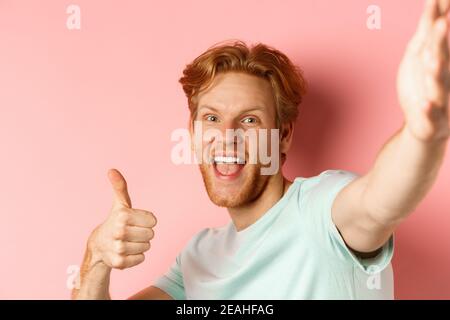 Aufgeregt Rotschopf Mann Tourist macht Selfie und zeigt Daumen-up, halten Kamera mit ausgestreckter Hand, stehend über rosa Hintergrund Stockfoto