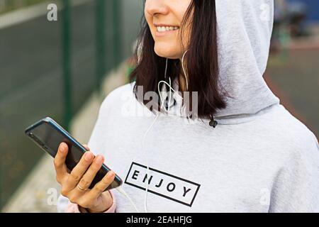 Eine kaukasische Frau nutzt ein Smartphone über kabelgebundene Kopfhörer mit einem Headset. Telefon in der Hand, weißer Draht im Ohr, genießen Etikett auf Hoodie. Hören Sie Musik über Stockfoto