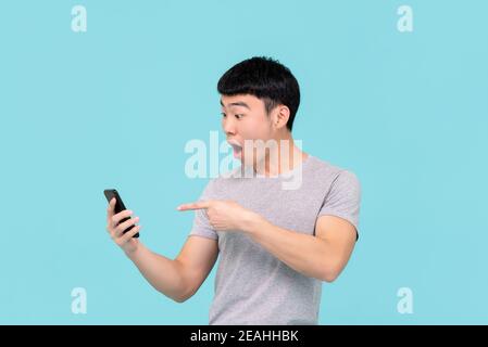 Schockiert aufgeregt junge asiatische Mann keuchend und zeigt auf mobile Telefon auf hellblauem Hintergrund isoliert Stockfoto