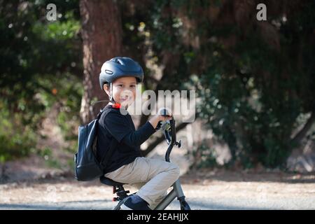 Kindergartenalter Junge mit einem großen Lächeln auf dem Fahrrad, trägt einen Rucksack und Helm zum Schutz. Stockfoto