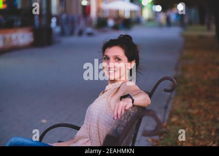 Porträt einer lächelnden jungen Frau. Elegante europäische Frau sitzt auf der Bank. Outdoor-Porträt von erstaunlichen weißen Mädchen Stockfoto