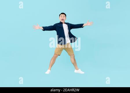 Ganzer Körper der jungen dynamischen schönen asiatischen Mann in casual Kleider suchen erstaunt und glücklich springen in der Luft mit Armen Und die Beine offen isoliert auf Licht b Stockfoto