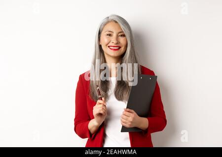 Erfolgreiche asiatische Dame Chef in roten Blazer, hält Zwischenablage mit Dokumenten und Stift, arbeiten und schauen glücklich, weißer Hintergrund Stockfoto