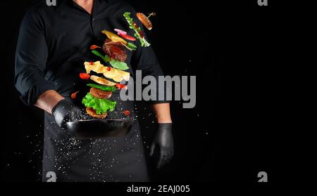 Mann in einer schwarzen Uniform, der einen Gusseisen um sich hält Bratpfanne mit schwebendem Cheeseburger Zutaten Stockfoto