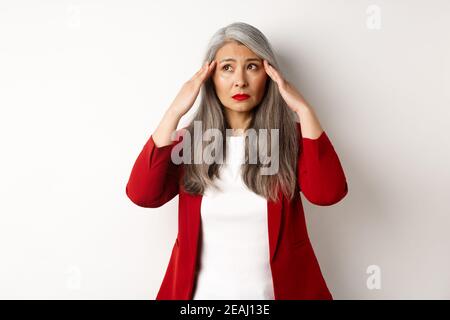 Unruhige asiatische Bürofrau, die den Kopf berührt, zur Seite schaut und alarmiert, versucht, sich zu beruhigen, über weißem Hintergrund stehend Stockfoto