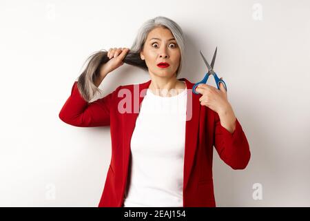 Asiatische ältere Frau hält Schere und Blick nervös an der Kamera, denken über das Schneiden von Haaren, Frisur ändern, stehen über weißem Hintergrund Stockfoto