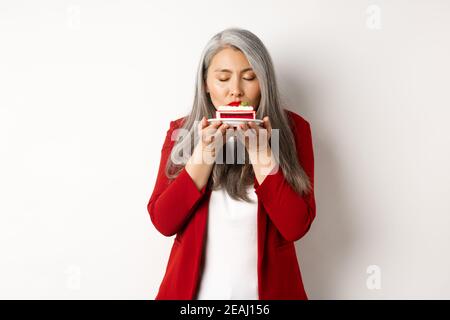 Bild von lustigen asiatischen Senior Dame schnüffeln leckeren Kuchen, Geruch Dessert mit zufriedenen Gesicht, über weißem Hintergrund stehen Stockfoto