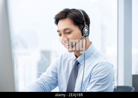 Lächelnd gut aussehende asiatische Mann arbeitet im Call Center Büro als Ein Kundendienstbetreiber Stockfoto