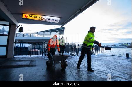 Hamburg, Deutschland. Februar 2021, 10th. Mitarbeiter eines Landschaftsbauunternehmens im Winterdienst verteilen auf den Landungsbrücken im Hafen Splitt. Im Hintergrund ist die markante Silhouette der Elbphilharmonie in der HafenCity zu sehen. Quelle: Christian Charisius/dpa/Alamy Live News Stockfoto