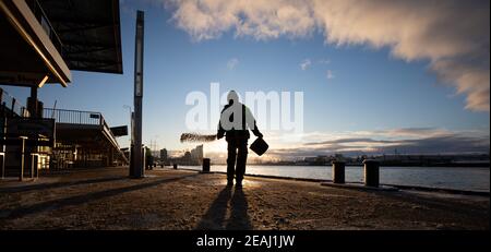 Hamburg, Deutschland. Februar 2021, 10th. Auf den Landungsbrücken im Hafen breitet sich ein Mitarbeiter eines Landschaftsbauunternehmens im Winterdienst Splitt aus. Im Hintergrund ist die markante Silhouette der Elbphilharmonie in der HafenCity zu sehen. Quelle: Christian Charisius/dpa/Alamy Live News Stockfoto