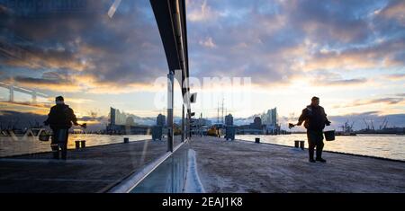 Hamburg, Deutschland. Februar 2021, 10th. Auf den Landungsbrücken im Hafen breitet sich ein Mitarbeiter eines Landschaftsbauunternehmens im Winterdienst Splitt aus. Im Hintergrund ist die markante Silhouette der Elbphilharmonie in der HafenCity zu sehen. Quelle: Christian Charisius/dpa/Alamy Live News Stockfoto