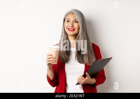 Stilvolle reife Geschäftsfrau in roten Blazer mit Pause, halten Zwischenablage und Kaffee trinken, Blick links mit zufrieden Lächeln, weißer Hintergrund Stockfoto