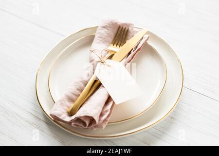 Goldene Rim Teller und goldenes Besteck mit leerem Anhänger auf weißem Tisch gebunden. Winkelansicht auf Tischeinstellung. Stockfoto