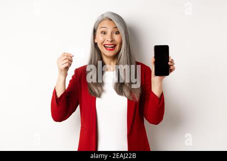 Fröhliche asiatische reife Frau zeigt leere Smartphone-Bildschirm und Kreditkarte, Konzept des E-Commerce Stockfoto