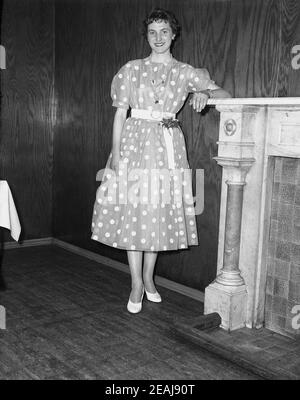 1950s, historisch, eine junge Dame mit einem gepunkteten oder gepunkteten Kleid, die in einem Veranstaltungsraum neben einem Kamin steht. Sie feiert ihren 21at. Geburtstag. Solch ein Partykleid mit Punkten auf einem Hintergrund mit einem Gürtel um die Mitte war in der Zeit des Retro Swing und Rockabiliy Ära, einer der frühen Stile der Rock and Roll Musik, in Mode. Stockfoto