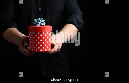 Erwachsener Mann in einem schwarzen Hemd hält in der Hand Eine runde rote Schachtel mit Punkten in blau Schleife Stockfoto