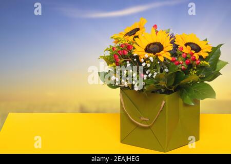 Nahaufnahme eines schönen Buketts mit gelben Sonnenblumen in einer dekorativen grünen Geschenkbox auf einem gelben Tisch auf einem verschwommenen blaugelben Hintergrund. Makro. Platz für Design. Kartenkonzept. Stockfoto