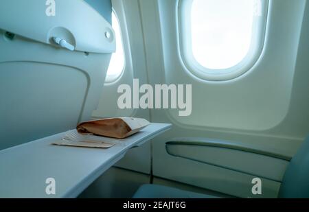 Ebenenfenster. Lebensmittel in brauner Papiertüte und Kunststoffgabel und Löffel auf Plastikflugzeug Tablett Tisch an der Sitzlehne. Fenster für Flugzeuge der Economy-Klasse. Innerhalb der kommerziellen Fluggesellschaft. Sitz mit Sessel. Stockfoto