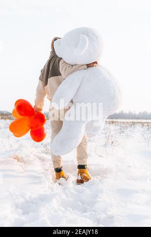 Junger Mann von hinten Umarmungen mit großen Teddybär weiches Spielzeug Geschenk Herz Luftballons an diesem besonderen Tag überraschende Freundin. Romantische Überraschung für Bi Stockfoto