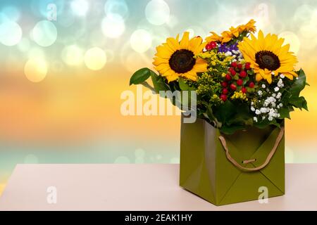 Nahaufnahme eines schönen Buketts mit gelben Sonnenblumen in einer dekorativen grünen Geschenkbox auf einem gelben Tisch auf einem verschwommenen blaugelben Hintergrund. Makro. Platz für Design. Kartenkonzept. Stockfoto