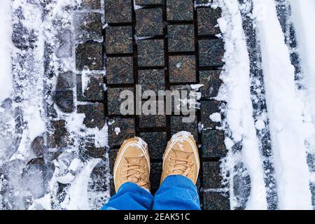 Steinweg im Park in der Schnee-Nahaufnahme. Damenfüße in Sneakers und Jeans auf einer verschneiten Piste, Draufsicht. Stockfoto