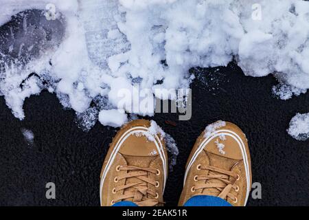 Steinweg im Park in der Schnee-Nahaufnahme. Damenfüße in Sneakers und Jeans auf einer verschneiten Piste, Draufsicht. Stockfoto