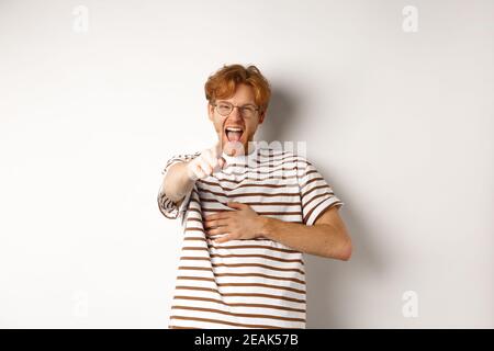 Junger Mann mit Ingwerhaar und Bart, der mit dem Finger auf die Kamera zeigt und lacht, macht sich lustig über jemanden, der auf weißem Hintergrund steht Stockfoto