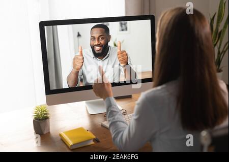 Videoanruf, Videokonferenz. Eine junge Frau diskutiert mit einem jungen afrikanischen Mann via Video einige Geschäftsaufgaben, sie sind zu einem Konsens und einer Show gekommen Stockfoto