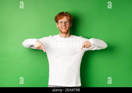 Gutaussehendes männliches Modell mit roten Haaren, Brille und Langarm tragend, Finger nach unten zeigend und fröhlich lächelnd, zeigt promo, grüner Hintergrund Stockfoto