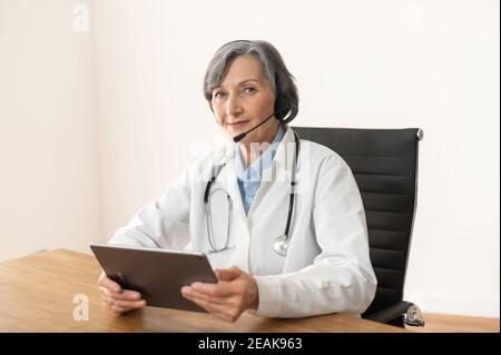 Ältere alte Ärztin mit Stethoskop in Headset und Laborkittel sitzen am Schreibtisch mit Touchpad, mit Online-Beratung mit Patienten oder Stockfoto