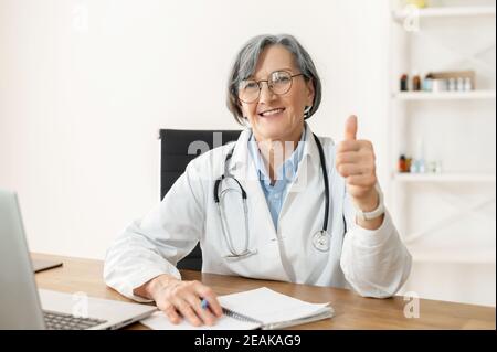 Positive ältere Ärztin mit einem Stethoskop in einem medizinischen Mantel sitzen am Schreibtisch mit einem Laptop, Notizen machen, zeigen einen Daumen-up, lächeln und Stockfoto