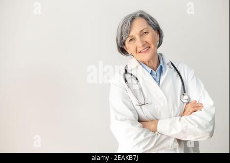 Positive ältere weibliche Ärztin mit einem Stethoskop in einem medizinischen Mantel posiert isoliert auf grauem Hintergrund, stehend mit Armen gefaltet, lächelnd und Stockfoto