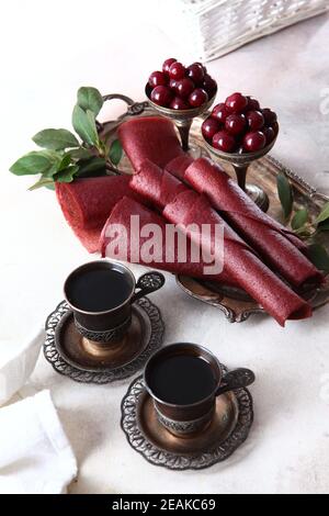 Kirschpastilien werden zu einem Kegel gerollt. Zwei Tassen Kaffee in Vintage-Geschirr. Vertikales Foto. Kirschen und Äpfel sind in einer Schüssel mit Nickel-Silber. Stockfoto